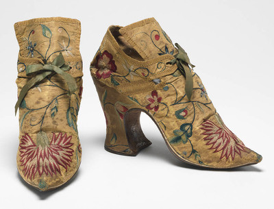 Textiles. Shoes/pumps. Silk taffeta, bound with ribbon; floss silk embroidery in French knots, long and short, satin and split stitches; tied with ribbon. Ground covered by a pattern of trailing and scrolling stems with leaves and flowers, carnation, rose, etc., so arranged that there is a carnation on each toe and a rose on each heel with a five petalled blue flower above it. The left shoe has a rose, the right a carnation at each side of the seam. Silk, length, whole, 7 in, height, back, 5.25 in, height, heel, 3 in, circa 1700-circa 1749. English.